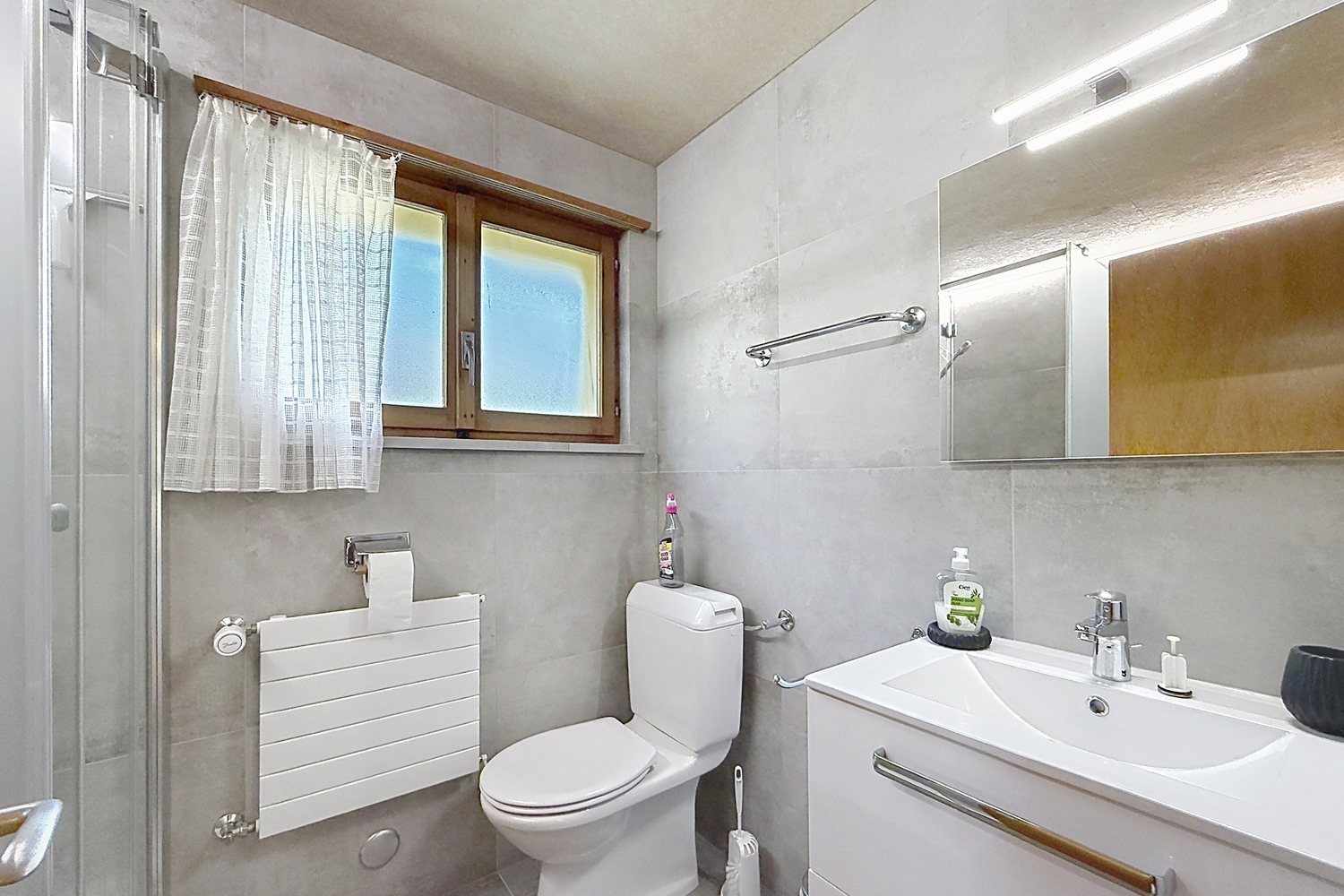 Salle de douche rénovée du premier étage : carrelage gris, douche vitrée, WC et meuble vasque blanc.