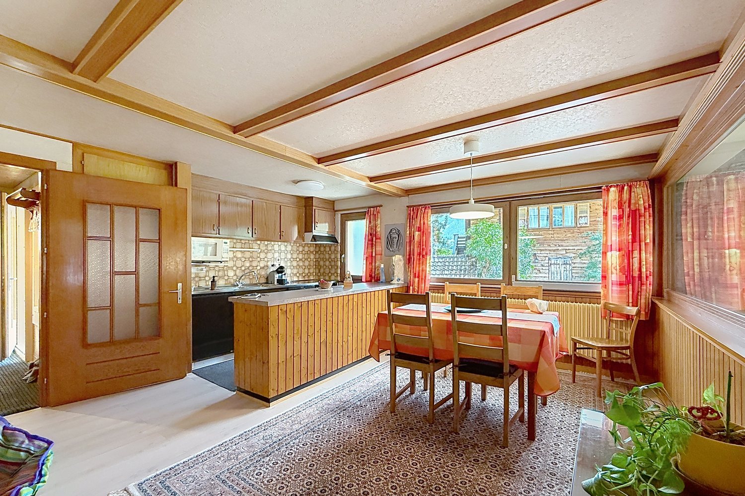 Séjour-salle à manger du premier étage sous un plafond à caissons bois, cuisine ouverte au fond et fenêtres sur le balcon.