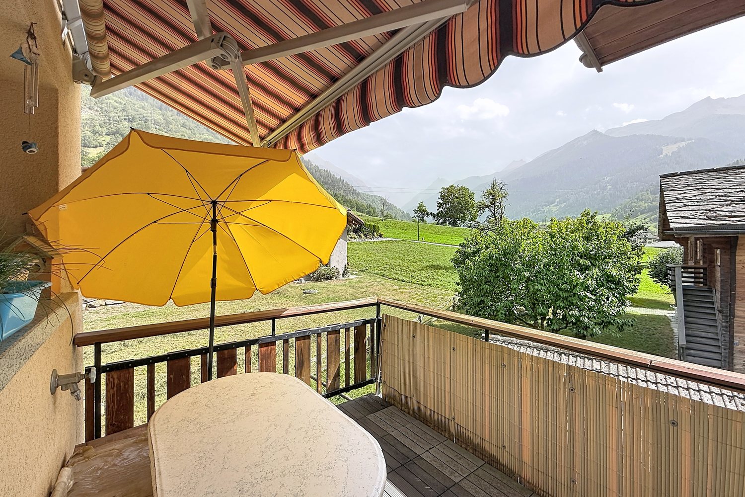 Balcon du duplex avec parasol jaune, store à rayures et vue sur les prés et les mayens de Bruson.