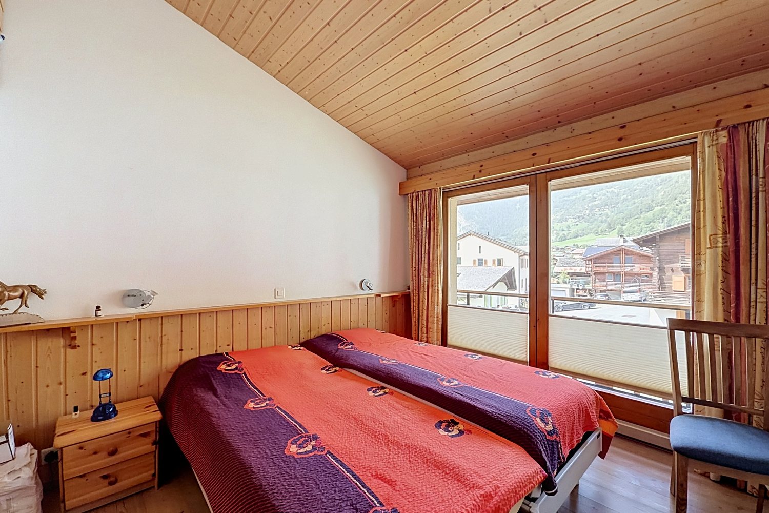Chambre du duplex sous un plafond lambrissé en pente, lit double et fenêtre avec vue sur le village.